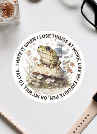 a sticker with a frog sitting on a mushroom surrounded by flowers, along with various office supplies such as a pair of glasses, a pen, and a notebook.