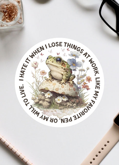 a sticker with a frog sitting on a mushroom surrounded by flowers, along with various office supplies such as a pair of glasses, a pen, and a notebook.