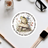 a sticker with a frog sitting on a mushroom surrounded by flowers, along with various office supplies such as a pair of glasses, a pen, and a notebook.