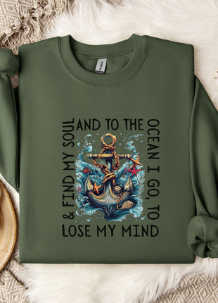a green sweatshirt with a graphic design of an anchor and waves, along with the text "& FIND MY SOUL AND TO THE OCEAN I GO TO LOSE MY MIND". The sweatshirt is laid out on a bed with a white blanket and a white mug.