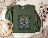 a green sweatshirt with a graphic design of an anchor and waves, along with the text "& FIND MY SOUL AND TO THE OCEAN I GO TO LOSE MY MIND". The sweatshirt is laid out on a bed with a white blanket and a white mug.