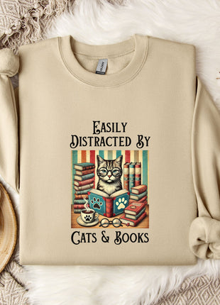a beige sweatshirt with a printed design of a cat surrounded by books and a cup of tea, along with the text "Easily Distracted By Cats & Books".