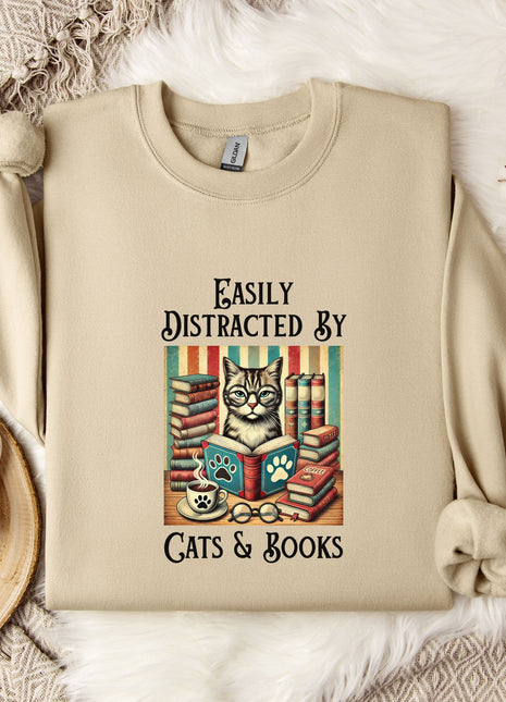 a beige sweatshirt with a printed design of a cat surrounded by books and a cup of tea, along with the text "Easily Distracted By Cats & Books".
