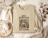 a beige sweatshirt with a printed design of a cat surrounded by books and a cup of tea, along with the text "Easily Distracted By Cats & Books".