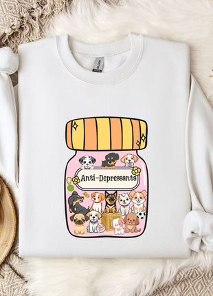 A white sweatshirt with a graphic design of a jar filled with various cartoon dogs and the text "Ani-Dependsents" printed on it. The sweatshirt is placed on a bed with a white blanket and a small white mug.
