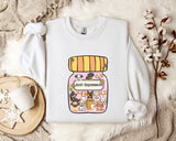 A white sweatshirt with a graphic design of a jar filled with various cartoon dogs and the text "Ani-Dependsents" printed on it. The sweatshirt is placed on a bed with a white blanket and a small white mug.