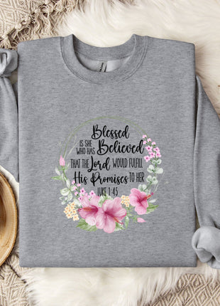 A gray sweatshirt with a floral design and text printed on it, placed on a bed with a white blanket and a white mug.