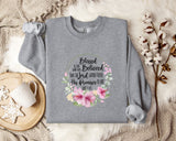 A gray sweatshirt with a floral design and text printed on it, placed on a bed with a white blanket and a white mug.
