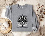 a gray sweatshirt with a graphic design on it, placed on a bed with a blanket and a mug.