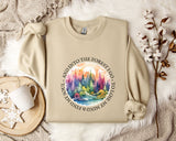 A beige sweatshirt with a colorful forest scene printed on it, along with a mug, a snowflake, and some cotton balls, all arranged on a white fur rug.