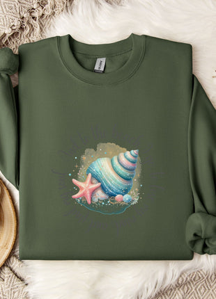 a green sweater with a colorful shell and starfish design, laid out on a white fur rug. Next to the sweater is a white mug with a heart-shaped handle, and a branch with white flowers.