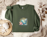 a green sweater with a colorful shell and starfish design, laid out on a white fur rug. Next to the sweater is a white mug with a heart-shaped handle, and a branch with white flowers.