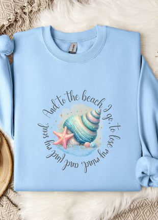 A light blue sweatshirt with a colorful shell and starfish design, along with a mug and a branch of cotton bolls, all placed on a white fur rug.