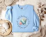 A light blue sweatshirt with a colorful shell and starfish design, along with a mug and a branch of cotton bolls, all placed on a white fur rug.