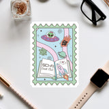 a colorful stamp with a whimsical illustration of a book, a rocket ship, and various other space-themed elements. The stamp is surrounded by various office supplies, including a pair of glasses, a pen, and a notebook.