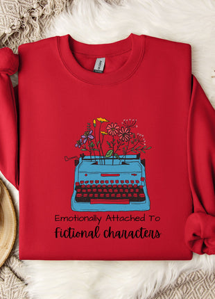 A red sweater with a graphic of a typewriter and flowers, along with the text "Emotionally Attached To Fictional Characters", is displayed on a bed with a white blanket and a white mug.