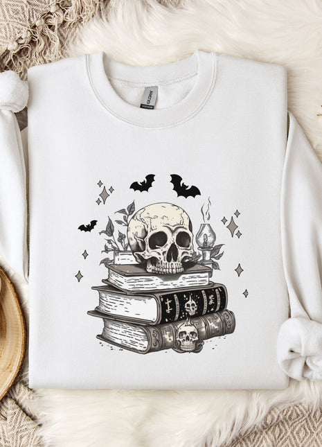 a white sweatshirt with a skull and bat design on it, along with a stack of books and some decorative elements such as a mug and a branch of cotton bolls.