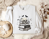 a white sweatshirt with a skull and bat design on it, along with a stack of books and some decorative elements such as a mug and a branch of cotton bolls.
