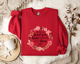 a red sweater with a floral design and text printed on it, placed on a white fur rug. There is also a white mug and a branch of cotton flowers nearby.