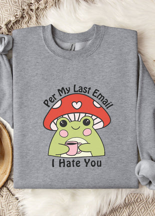 a gray sweatshirt with a cartoon frog wearing a red mushroom hat and the text "Per My Last Email I Hate You" printed on it. The sweatshirt is laid out on a bed with a blanket and a mug, and there are some white flowers nearby.