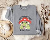 a gray sweatshirt with a cartoon frog wearing a red mushroom hat and the text "Per My Last Email I Hate You" printed on it. The sweatshirt is laid out on a bed with a blanket and a mug, and there are some white flowers nearby.
