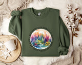 a green sweatshirt with a colorful forest scene printed on it, along with a mug and some white flowers on a bed.