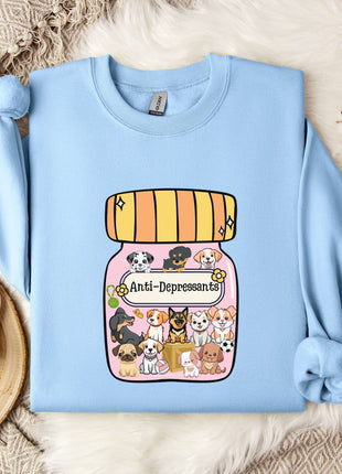 A light blue sweatshirt with a graphic of a jar filled with cartoon dogs and the text "Anti-Depressants" printed on it, along with a mug and some flowers on a bed.