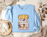 A light blue sweatshirt with a graphic of a jar filled with cartoon dogs and the text "Anti-Depressants" printed on it, along with a mug and some flowers on a bed.