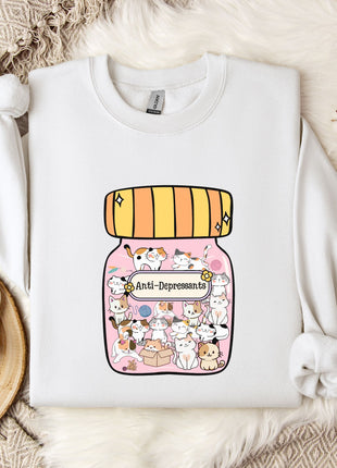 A white sweatshirt with a graphic design of a jar filled with various cartoon animals, including cats and dogs, and the text "Anti-Depressants" printed on it. The sweatshirt is laid out on a bed with a white blanket and a small white mug.