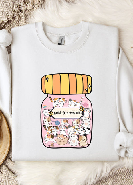 A white sweatshirt with a graphic design of a jar filled with various cartoon animals, including cats and dogs, and the text "Anti-Depressants" printed on it. The sweatshirt is laid out on a bed with a white blanket and a small white mug.