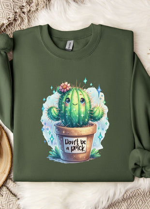 a green sweatshirt with a printed design of a cactus and the text "Don't be a brick" on it. The sweatshirt is laid out on a bed with a white blanket and a white mug. There are also some white flowers and a white branch visible in the background.