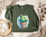 a green sweatshirt with a printed design of a cactus and the text "Don't be a brick" on it. The sweatshirt is laid out on a bed with a white blanket and a white mug. There are also some white flowers and a white branch visible in the background.