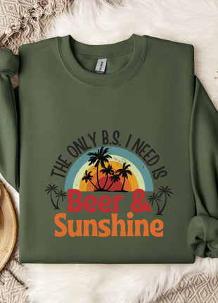 A green sweatshirt with a graphic design that says "The only B.S. I need is Beer & Sunshine" printed on it, along with a palm tree and sunset. The sweatshirt is laid out on a white fur rug, and there is a mug and some flowers nearby.