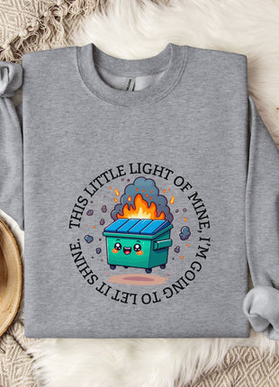 a gray sweatshirt with a graphic design on it, including a cartoon image of a fire-breathing trash can. The sweatshirt is laid out on a bed with a blanket and a mug, and there are some cotton balls and a plant nearby.