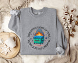 a gray sweatshirt with a graphic design on it, including a cartoon image of a fire-breathing trash can. The sweatshirt is laid out on a bed with a blanket and a mug, and there are some cotton balls and a plant nearby.