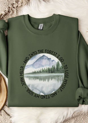a green sweatshirt with a circular design on it, depicting a mountain landscape with a lake and pine trees. The design is surrounded by the text "And into the forest I go - to lose my self and find my soul".
