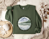 a green sweatshirt with a circular design on it, depicting a mountain landscape with a lake and pine trees. The design is surrounded by the text "And into the forest I go - to lose my self and find my soul".