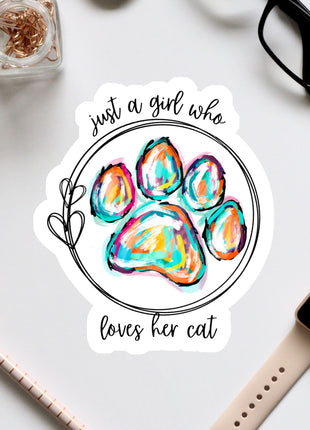 A sticker with a paw print and the text "Just a girl who loves her cat" is surrounded by various office supplies, including a pair of glasses, a pen, and a watch.