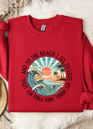 A red sweater with a graphic design of a beach scene and the text "To the beach I go, I lose my soul, and I lose my mind" printed on it, along with a palm tree and ocean waves. The sweater is laid out on a white fur rug, and there is a white mug and a branch of cotton flowers nearby.