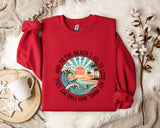 A red sweater with a graphic design of a beach scene and the text "To the beach I go, I lose my soul, and I lose my mind" printed on it, along with a palm tree and ocean waves. The sweater is laid out on a white fur rug, and there is a white mug and a branch of cotton flowers nearby.