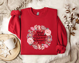 A red sweater with a floral design and text printed on it, along with a mug, a white mug, and some white flowers on a white fur rug.