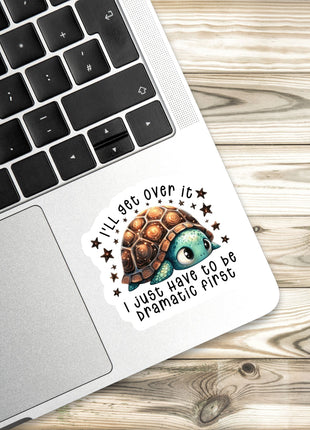 A sticker with a cartoon turtle and the text "I'll get over it, I just have to be dramatic first" is placed on a laptop keyboard.