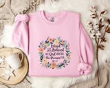 A pink sweatshirt with a floral design and text, placed on a white fur rug next to a white mug and a branch of cotton bolls.