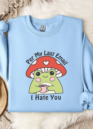 a light blue sweatshirt with a cartoon frog wearing a red mushroom hat and the text "Per My Last Email I Hate You" printed on it. The sweatshirt is laid out on a bed with a white blanket and a mug, and there are some white flowers in the background.