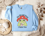 a light blue sweatshirt with a cartoon frog wearing a red mushroom hat and the text "Per My Last Email I Hate You" printed on it. The sweatshirt is laid out on a bed with a white blanket and a mug, and there are some white flowers in the background.