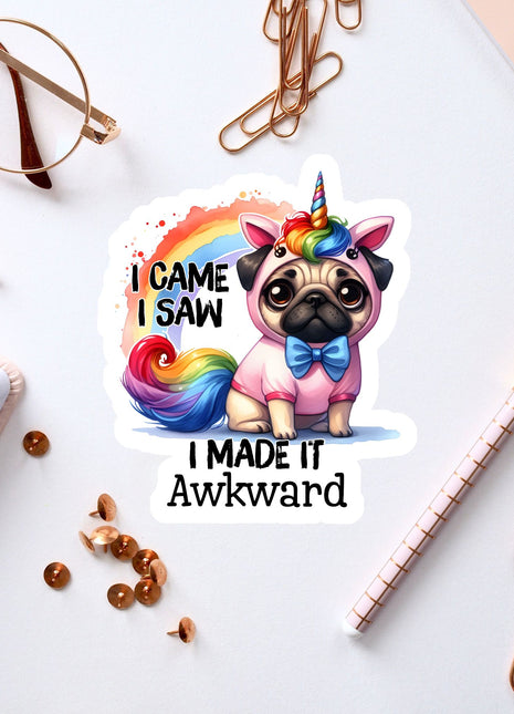 A sticker featuring a pug dog wearing a unicorn costume, with the text "I CAME I SAW I MADE IT AWKWARD" printed on it. The sticker is surrounded by various office supplies, such as a pen, a pair of glasses, and a notebook.