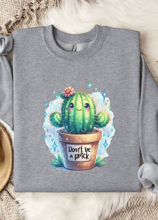 a gray sweatshirt with a colorful illustration of a cactus and the text "Don't be a prick" printed on it. The sweatshirt is laid out on a bed with a white blanket and a mug, and there are some cotton balls and a plant in a pot nearby.