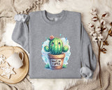 a gray sweatshirt with a colorful illustration of a cactus and the text "Don't be a prick" printed on it. The sweatshirt is laid out on a bed with a white blanket and a mug, and there are some cotton balls and a plant in a pot nearby.