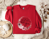 a red sweater with a floral design and text printed on it, placed on a white fur rug. There is also a white mug and a branch of cotton flowers nearby.