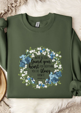 a green sweatshirt with a floral wreath design and a quote printed on it, placed on a bed with a white blanket and a white mug.